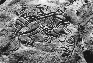 Gravure rupestre représentant un bovin, deux personnages lui installent une charge sur les cornes et un troisième lui tend une auge